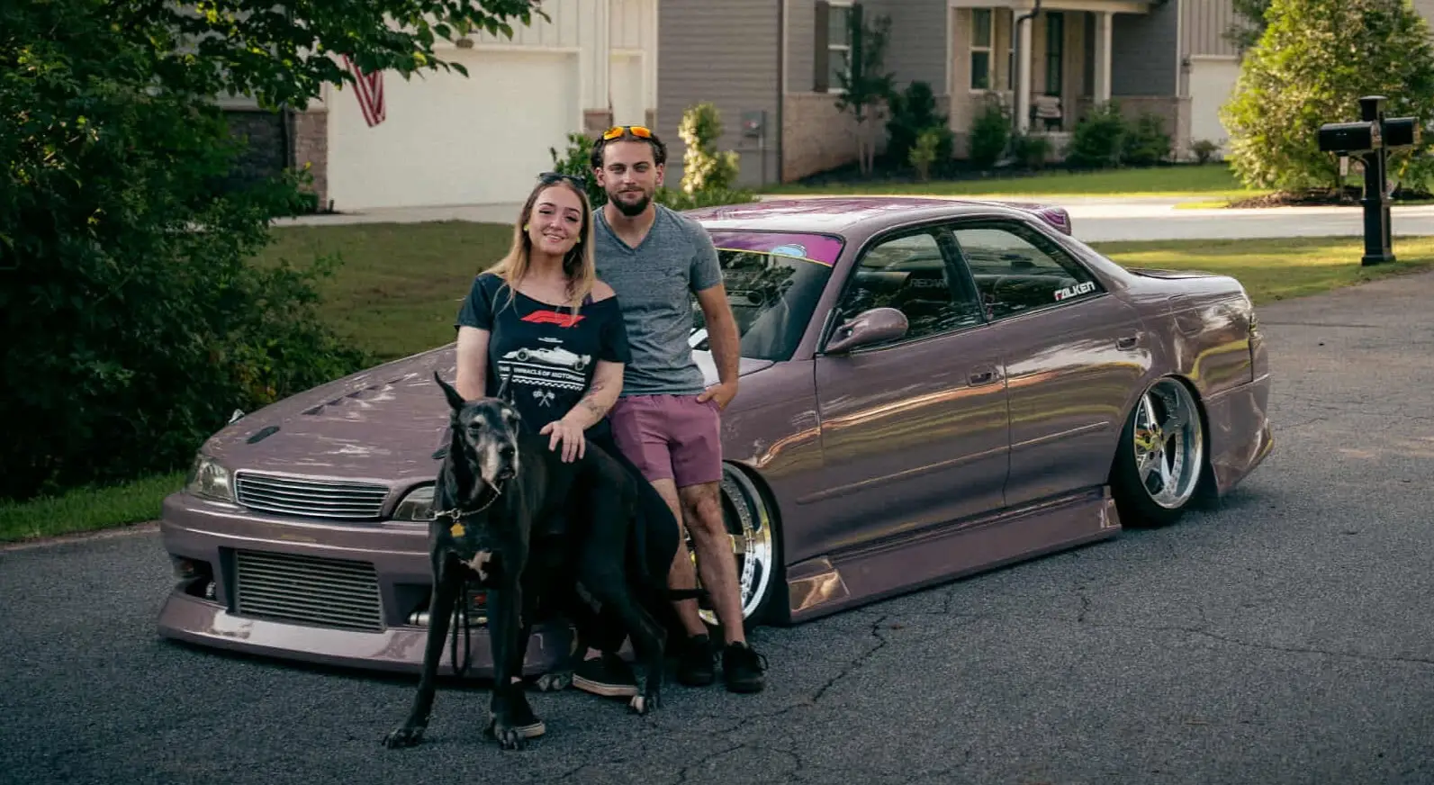 Jacob Gerken and family with their Great Dane beside a JZX90 Flatout Manufacturing About Section Blonde women and brunette man standing with their large black Great Dane in front of a pink JZX90 (car).